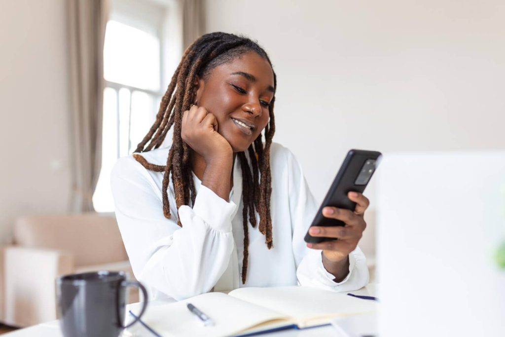 Young smiling African business woman using smartphone near computer in office without understanding attention residue