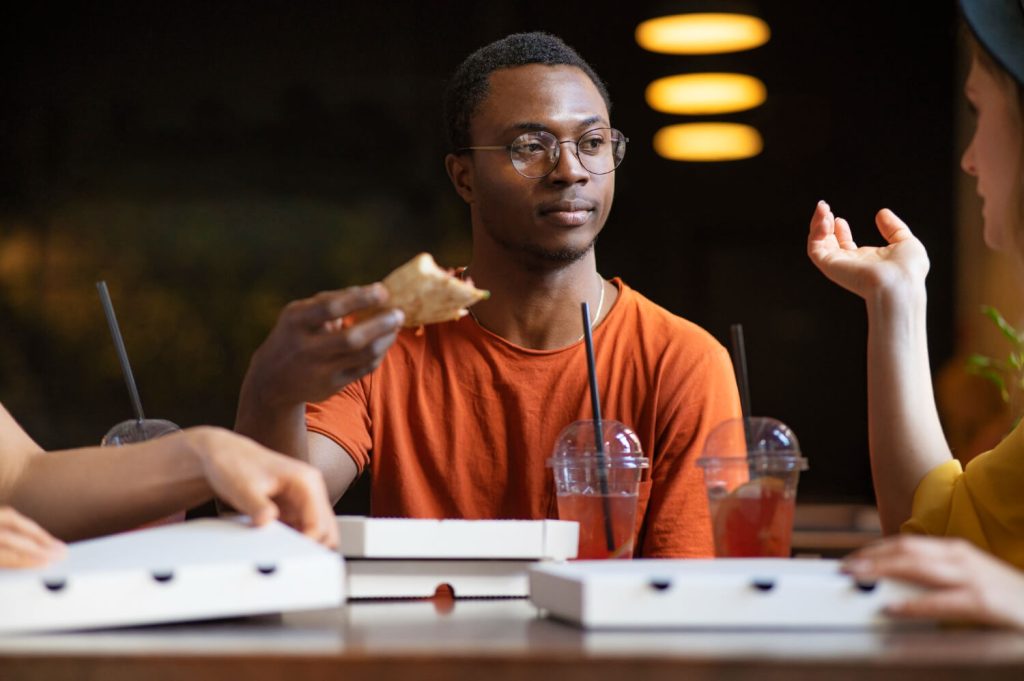 young black man eating pizza and enjoying it whereas his friends don't depicting the subjectivity of experience and why experience is the best teacher is not always true