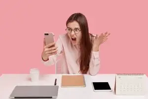 Attractive female blogger struggling with perfectionism clenches teeth, holds brocken tablet, cant understand why it doesnt work, has many things neatly arranged on desk, drinks coffee