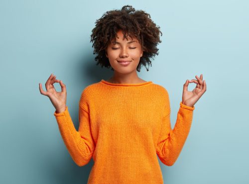 fro American woman meditates indoor, keeps hands in mudra gesture, has eyes closed as she seeks to make mindful choices